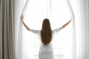 A Person In A White Robe Stands Facing A Window, Holding Open White Curtains, Letting In Bright Natural Light.