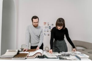 Two People Stand At A Table Reviewing Fabric Samples And Design Materials In A Light, Modern Room.