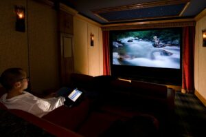 Person Sitting In A Home Theater Watching A Nature Scene On A Large Screen, Holding A Tablet Displaying A Bar Graph.