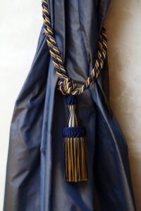 Dark Blue Curtain Tied With A Gold And Blue Striped Tassel Tieback Against A Light Colored Wall.
