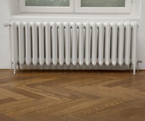 White Radiator Positioned Against A Wall Beneath A Window, With Wooden Herringbone Patterned Flooring.