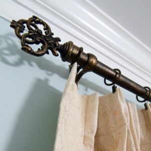 An Ornate Bronze Curtain Rod With A Decorative Finial Holds Beige Patterned Curtains Against A Light Blue Wall Near A White Ceiling.