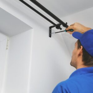 Person In A Blue Uniform Installs A Black Curtain Rod On A White Wall Using A Screwdriver.