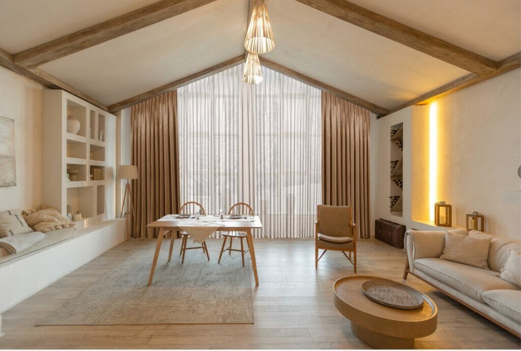 Cozy living room with wooden ceiling, a dining table for two, a couch, shelves, and floor-to-ceiling curtains. Soft lighting illuminates the space.