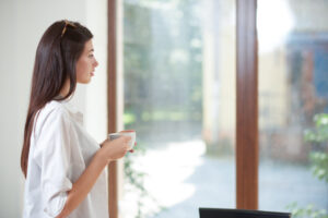 Woman In A White Shirt Holds A Mug And Looks Out A Window, With Greenery Visible Outside On A Sunny Day.