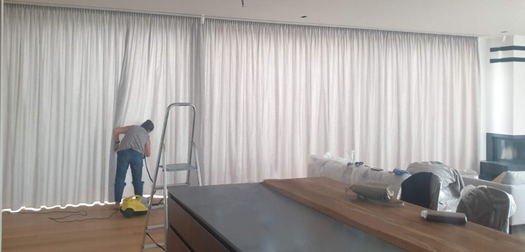 A person vacuuming custom curtains in a modern living room with a ladder nearby and furniture partially covered with sheets.