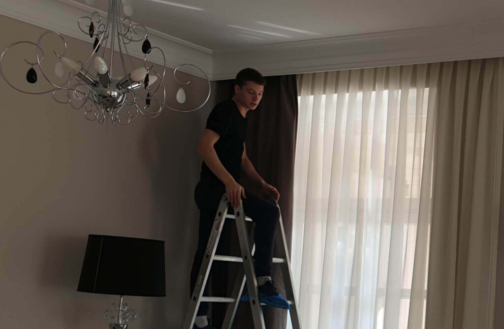Man on a ladder under a chandelier looking toward newly installed custom curtains.